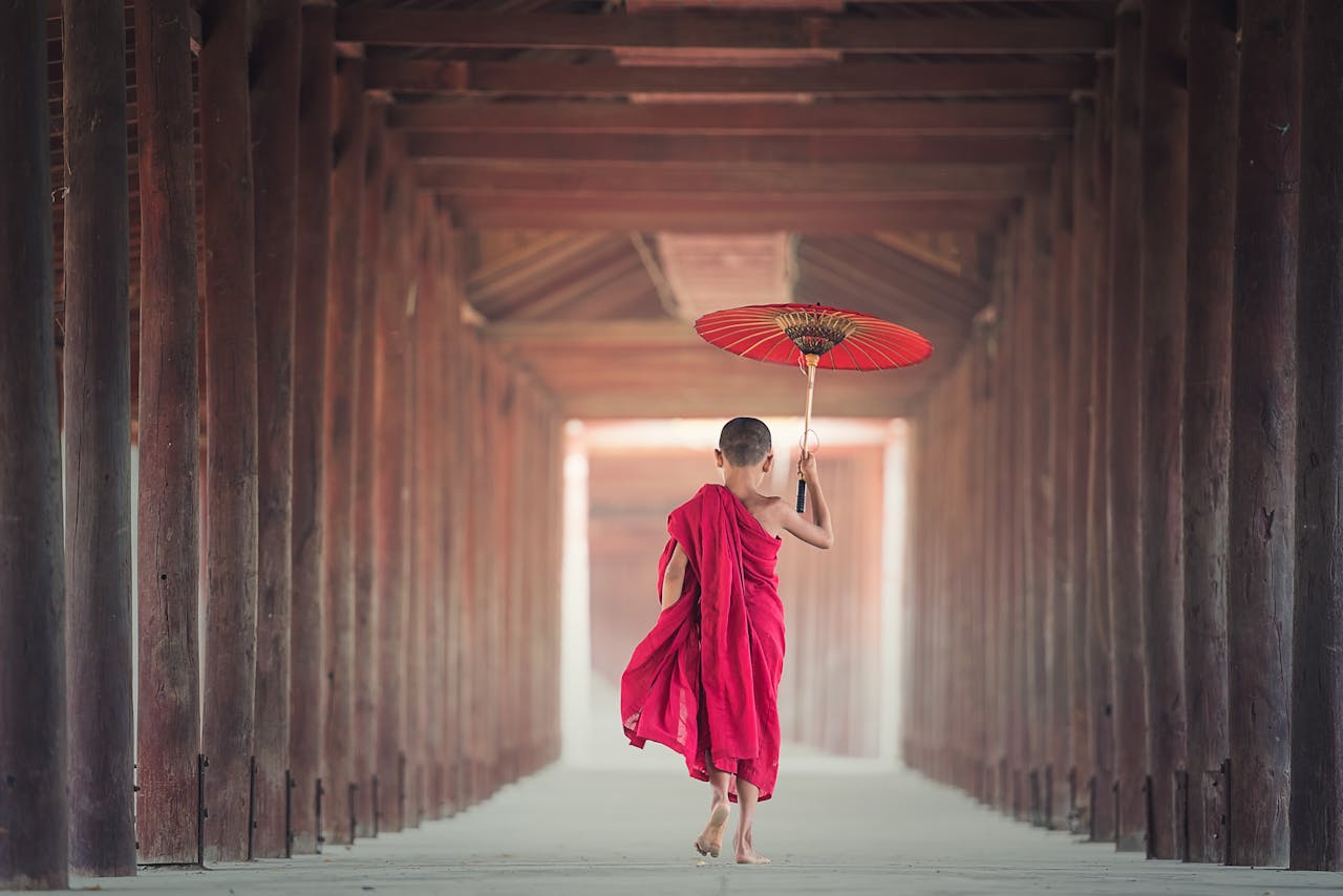 A child monk holding a parasol walks through a traditional Asian temple corridor, embracing cultural heritage.