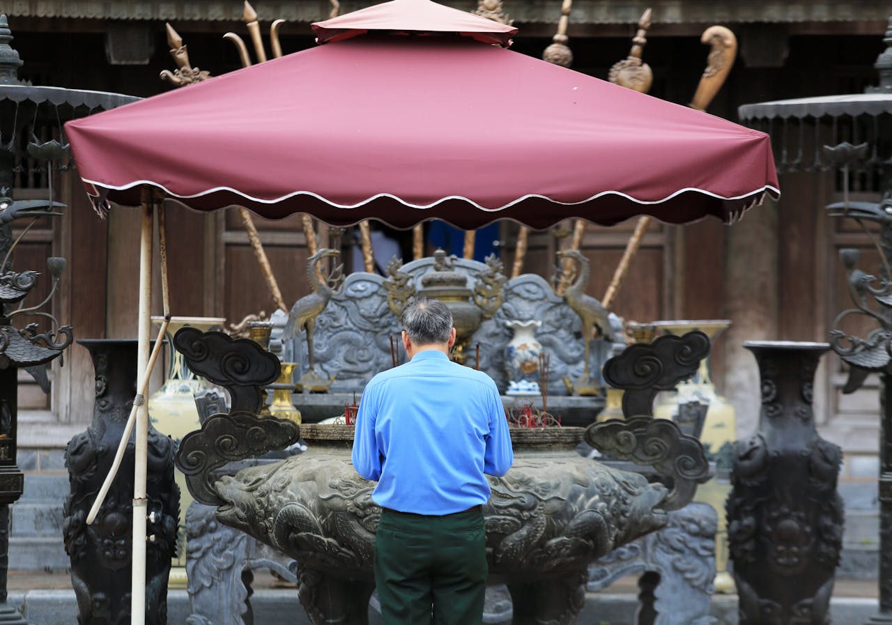 An adult man prays at an Asian temple under a red canopy, surrounded by ornate decor.
